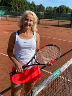 Frau mit halblangen weißen Haaren im weißen Tennisdress mit rotem Rock steht lächelnd mit dem Tennisschläger in der Hand am Tennisnetz
